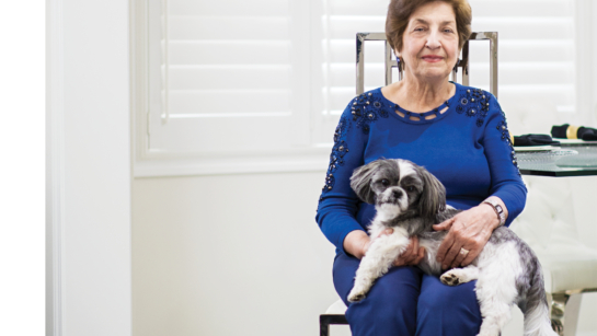 Shamoon Amir Nasir sitting in a bright white living room, she has a small dog in her lap and is smiling.