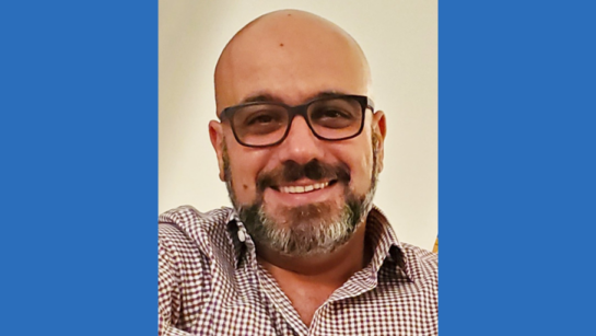 Dr. Koorosh Shirkool, a Krembil Brain Institute neurologist, smiles for a selfie while wearing a checkered collared shirt and dark framed glasses in front of a yellow wall.