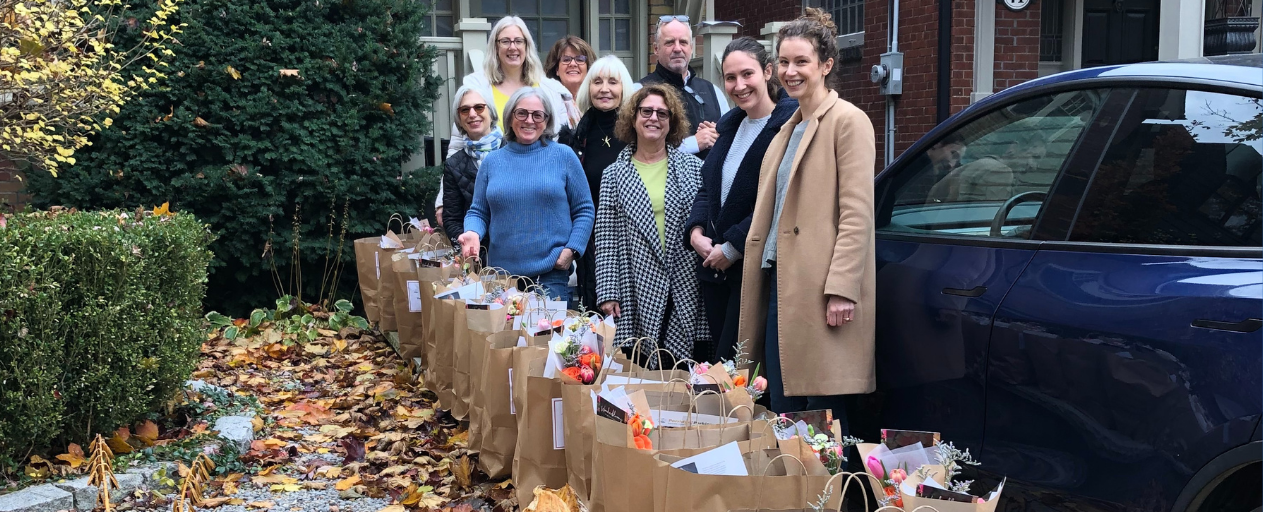 A group of volunteers stand in a drive way on a autumn day with rows of paper bags filled with food for delivery