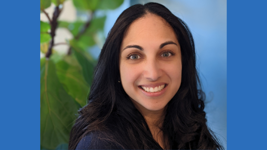 Marsha Alvares looks into the camera and smiles with a blue background and plant behind her.