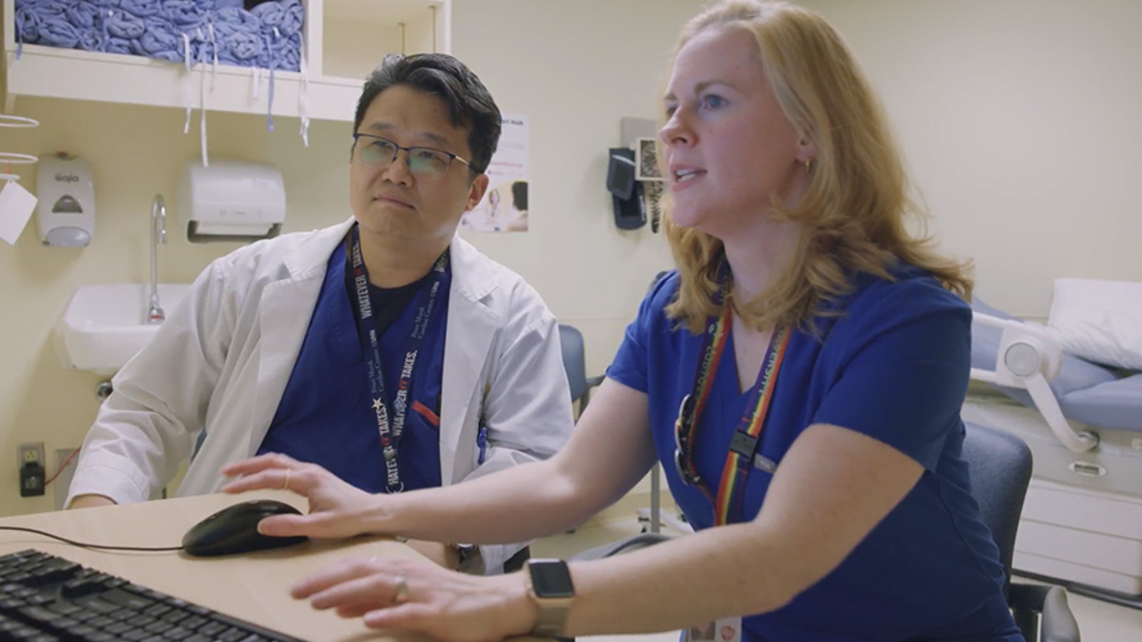 Two healthcare professionals, one wearing glasses and a lanyard and the other in blue scrubs, sitting at a desk and looking at a computer screen in a hospital setting.