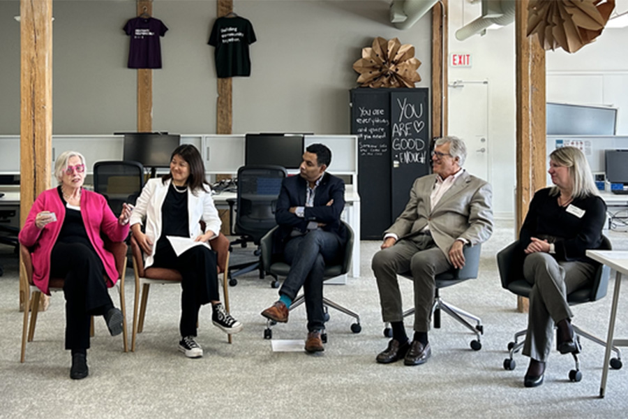 Five individuals seated in a row, participating in a panel discussion in a casual meeting room with a motivational quote on a blackboard behind them. From left to right: the first person is gesturing with their hand while speaking, the next three are attentively listening, and the last person holds a paper, possibly to take notes or ask questions. On the wall behind, there are several t-shirts displayed on hangers, and a sign that reads "You are good enough".