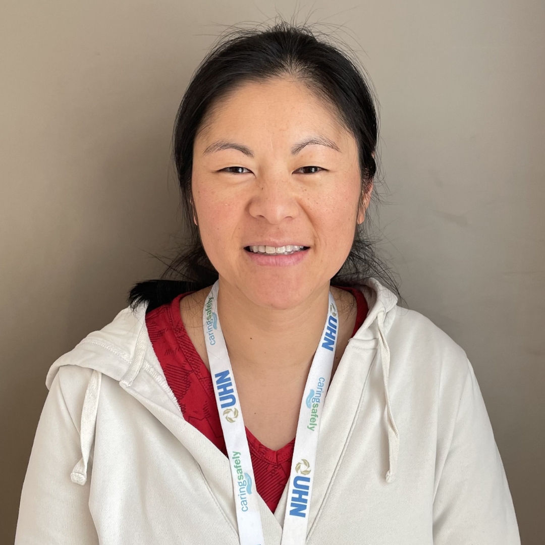 Vanessa Ong with a smile wearing a white hoodie and a lanyard with visible text that reads "UHN." The background is a plain, light tan color.