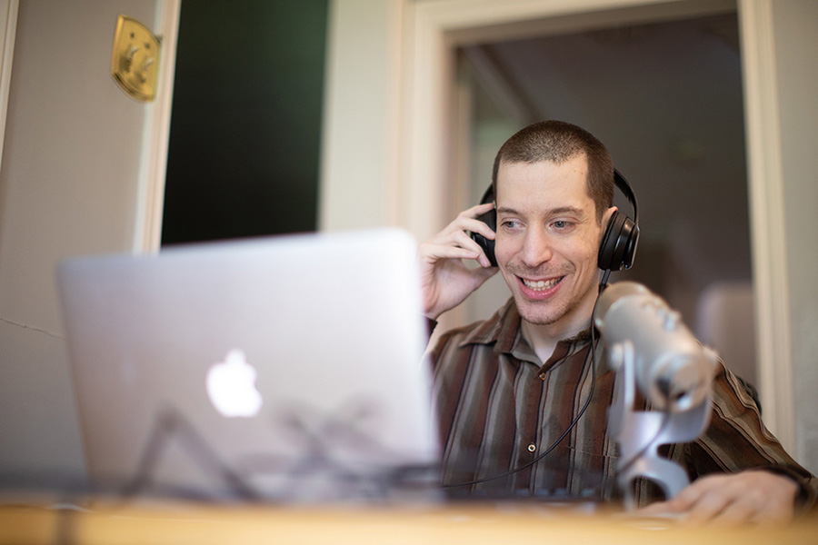 Person using an Apple laptop and speaking on a mobile phone while wearing headphones, seated at a desk with a microphone in front of them. The individual appears to be engaged in a conversation and smiling.