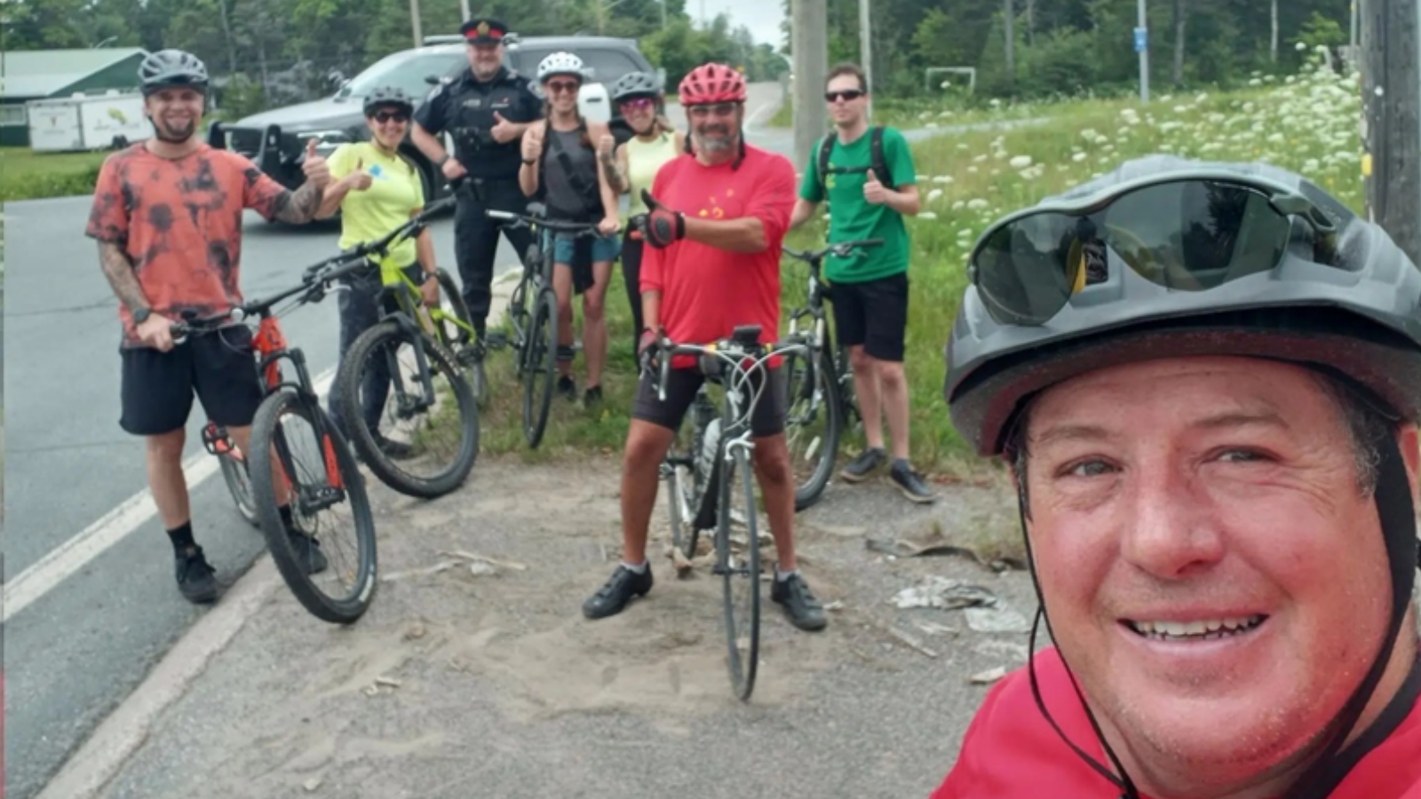 Group of seven cyclists in various outfits with helmets, posing on a roadside; a mix of mountain bikes and road bikes are visible. Lush greenery and wildflowers in the background, with a clear sky. Closest to the foreground, a smiling person with a helmet taking a selfie.