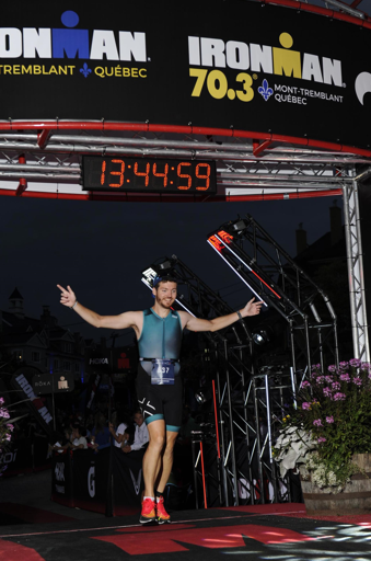 Troy Wagner wearing a blue triathlon suit and cap, marked with the number 57, celebrates as they cross the finish line at the IRONMAN 70.3 Mont-Tremblant, Quebec. The digital clock overhead displays the time 13:44:59.
