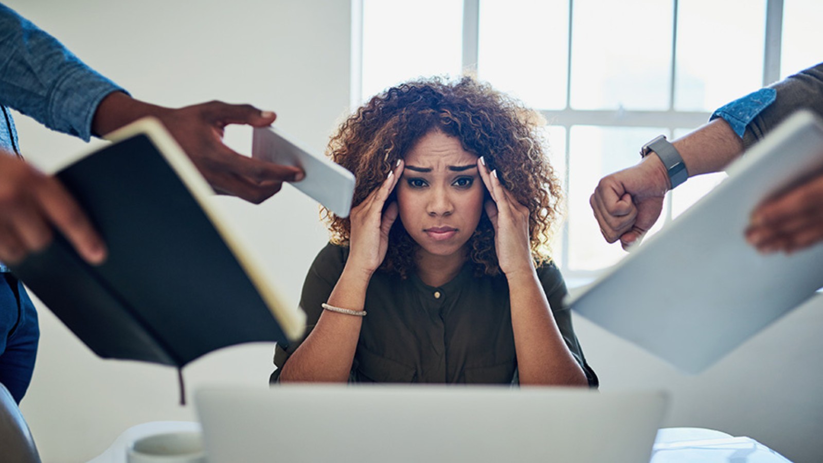 A woman holding her head while at an office with others requesting things of her.