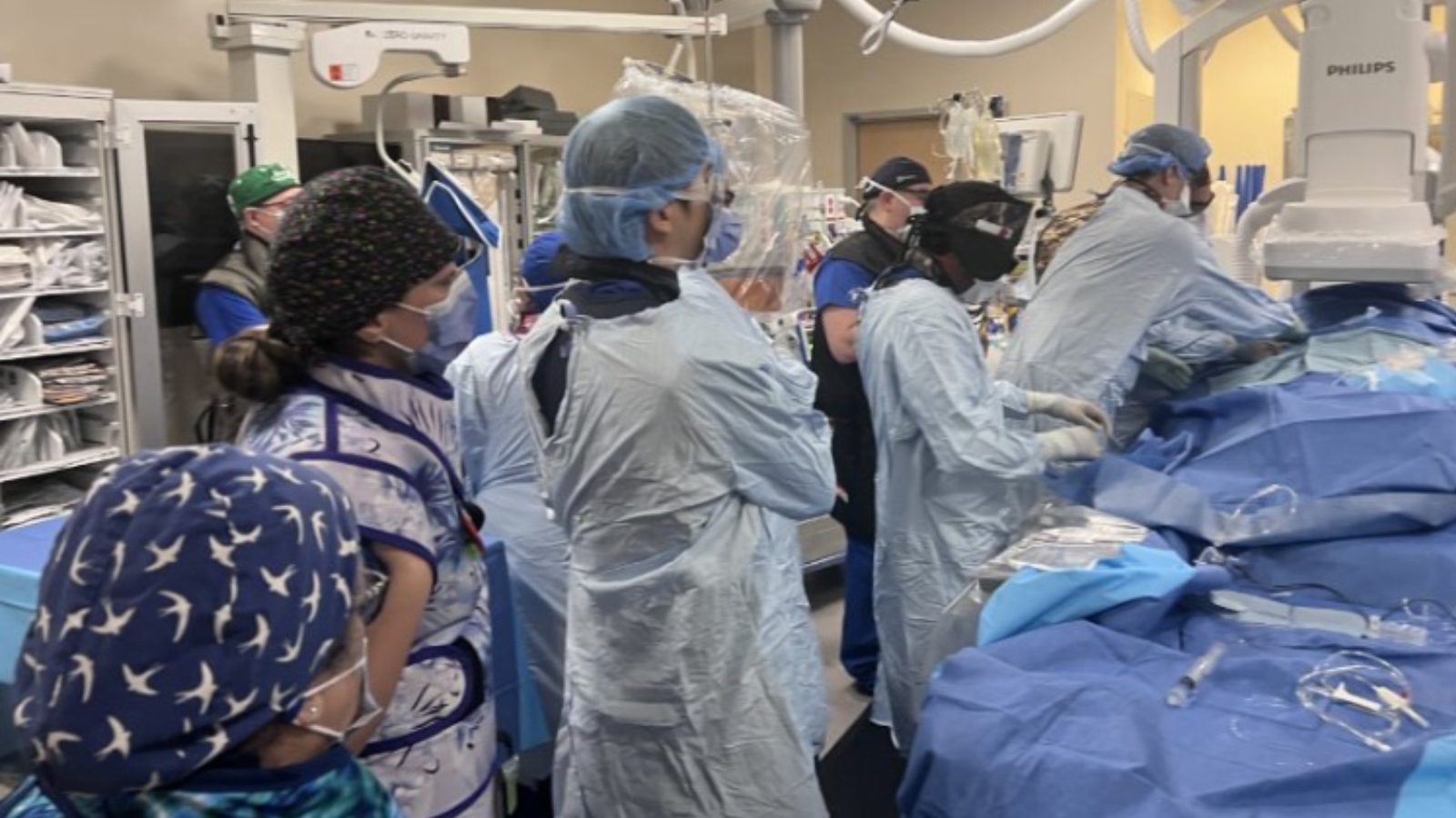 A group of medical professionals in scrubs and surgical gowns are gathered around a patient undergoing surgery in a hospital operating room equipped with advanced medical equipment.