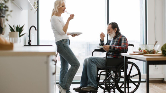 A photo of a woman standing and eating while leaning against a kitchen counter. A man is seated in a wheelchair next to her eating. A bright window is in the background with lots of light shining in.