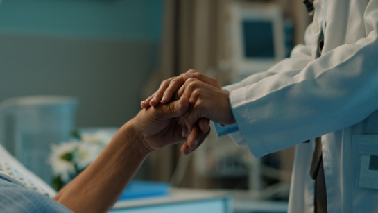 A stock image of a hospital bedside with a patient and doctor holding hands